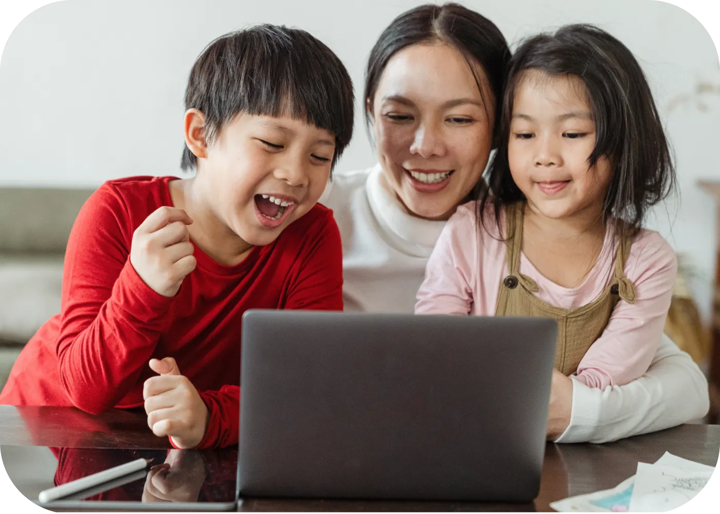 A boy pumps his fist in delight in front of a laptop during an online lesson, while his mother and little sister smile beside him